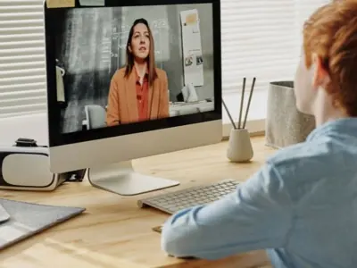 A boy writes in front of a computer with a woman speaking on the screen